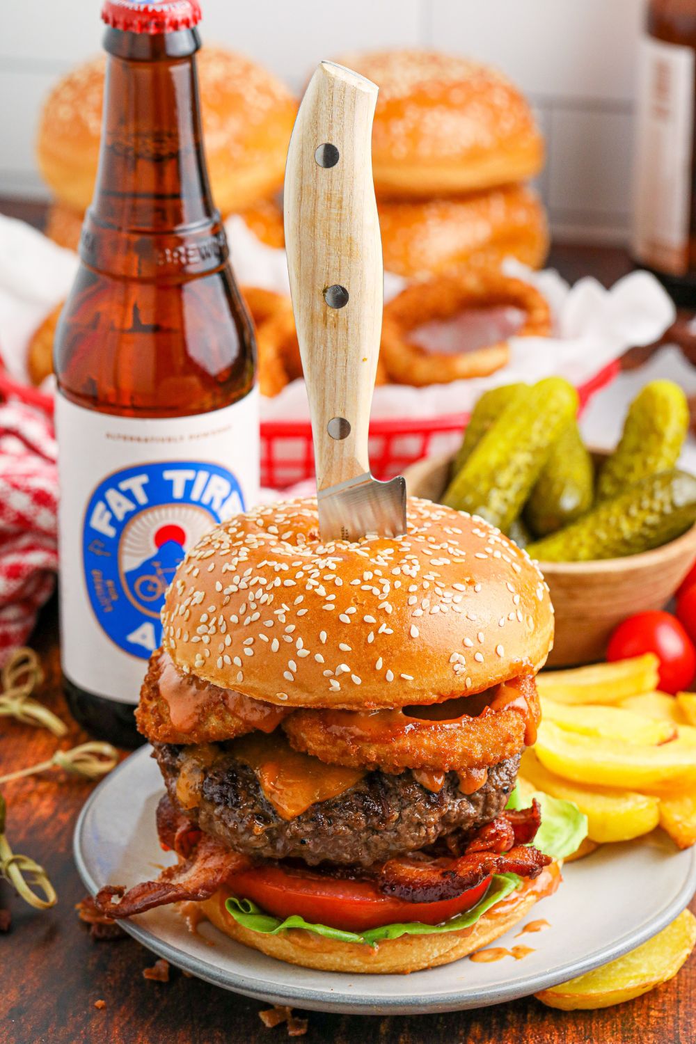 Cowboy Burger with a knife stabbed in center of bun with pickles and beer bottle in background
