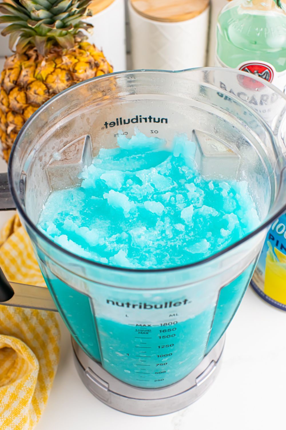 Frozen Blue Hawaiian Cocktail in a blender with ingredients for drink behind it on table 