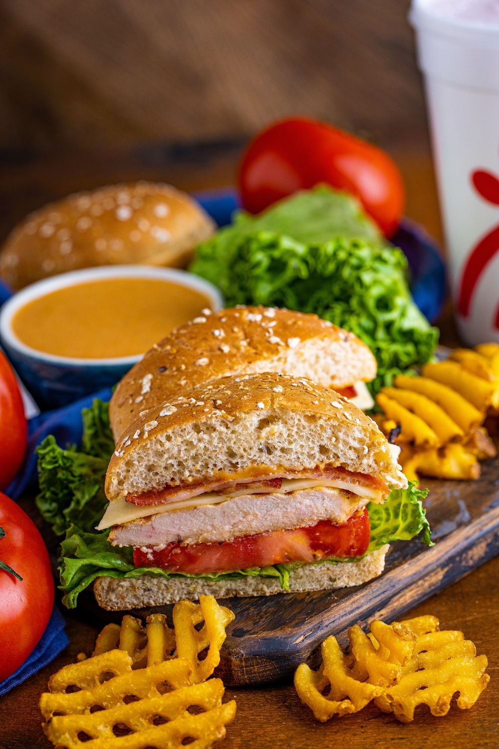 Chick Fil A Grilled Chicken Sandwich sliced in half sitting on table with lettuce and tomatoes around the sandwich and a dipping sauce 