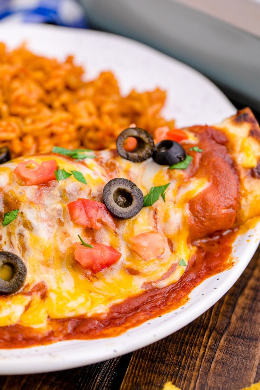 cheesy enchilada on plate covered in sauce and cheese topped with tomatoes, olives, and rice to side