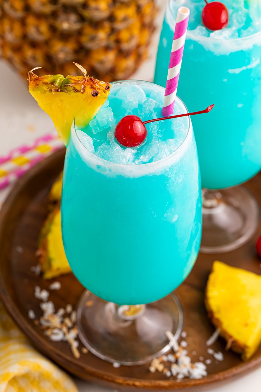 Frozen Blue Hawaiian Cocktails on table with a close up of class to see the cherry in center of drink and pineapple wedge on side of glass 