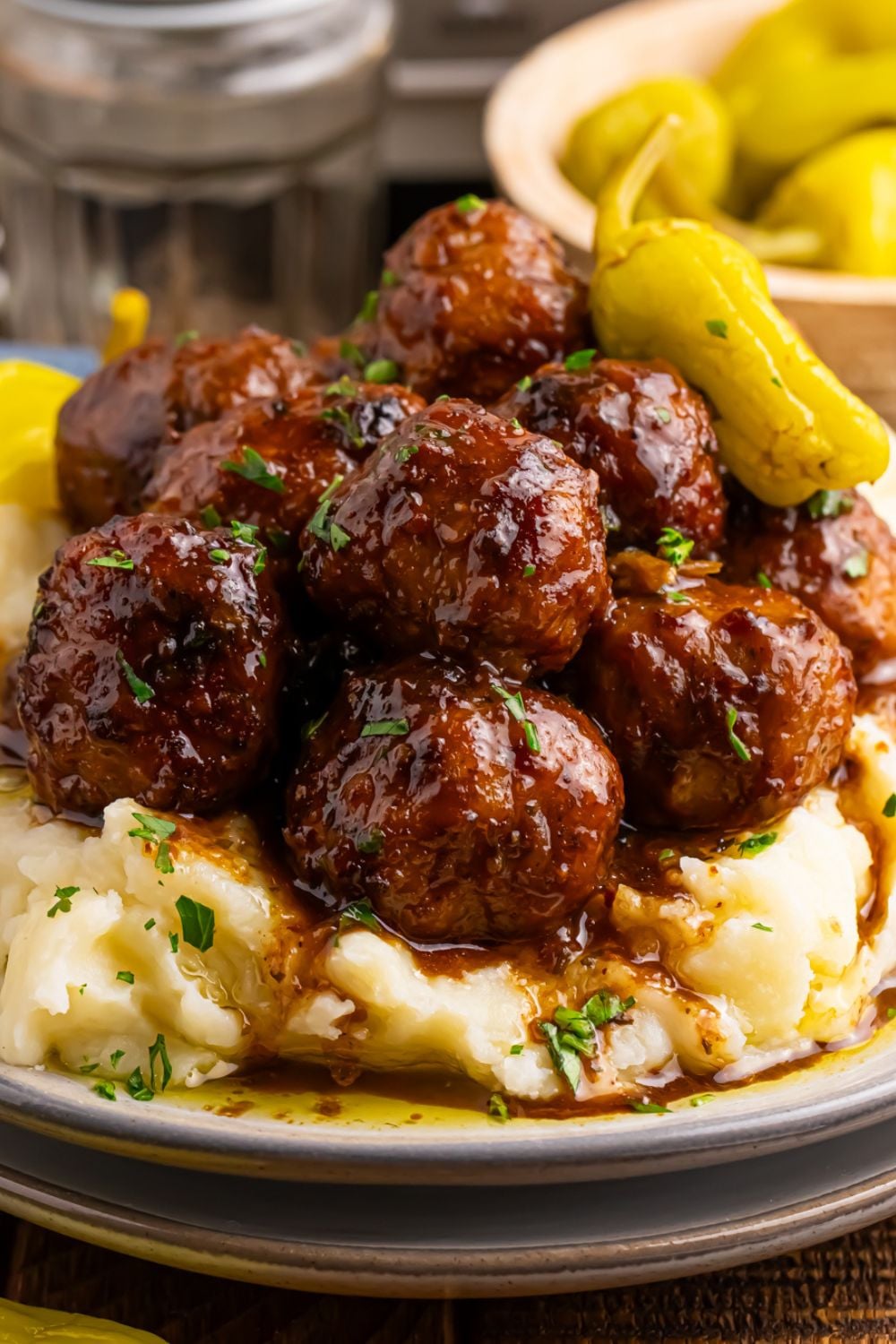 Crockpot Mississippi Meatballs piled in a stack on a plate with a bowl of peppers behind it on table and mashed potatoes on the plate with meatballs