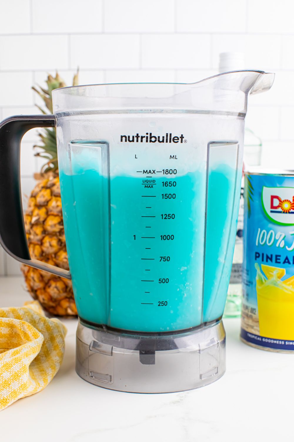 side view of Frozen Blue Hawaiian Cocktail in a blender on table with pineapple juice and whole pineapple behind it on table