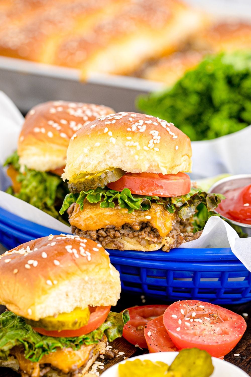 Classic Cheeseburger Sliders in baskets and spread out on table with tomatoes and ketchup around the basket, and a bowl of pickles