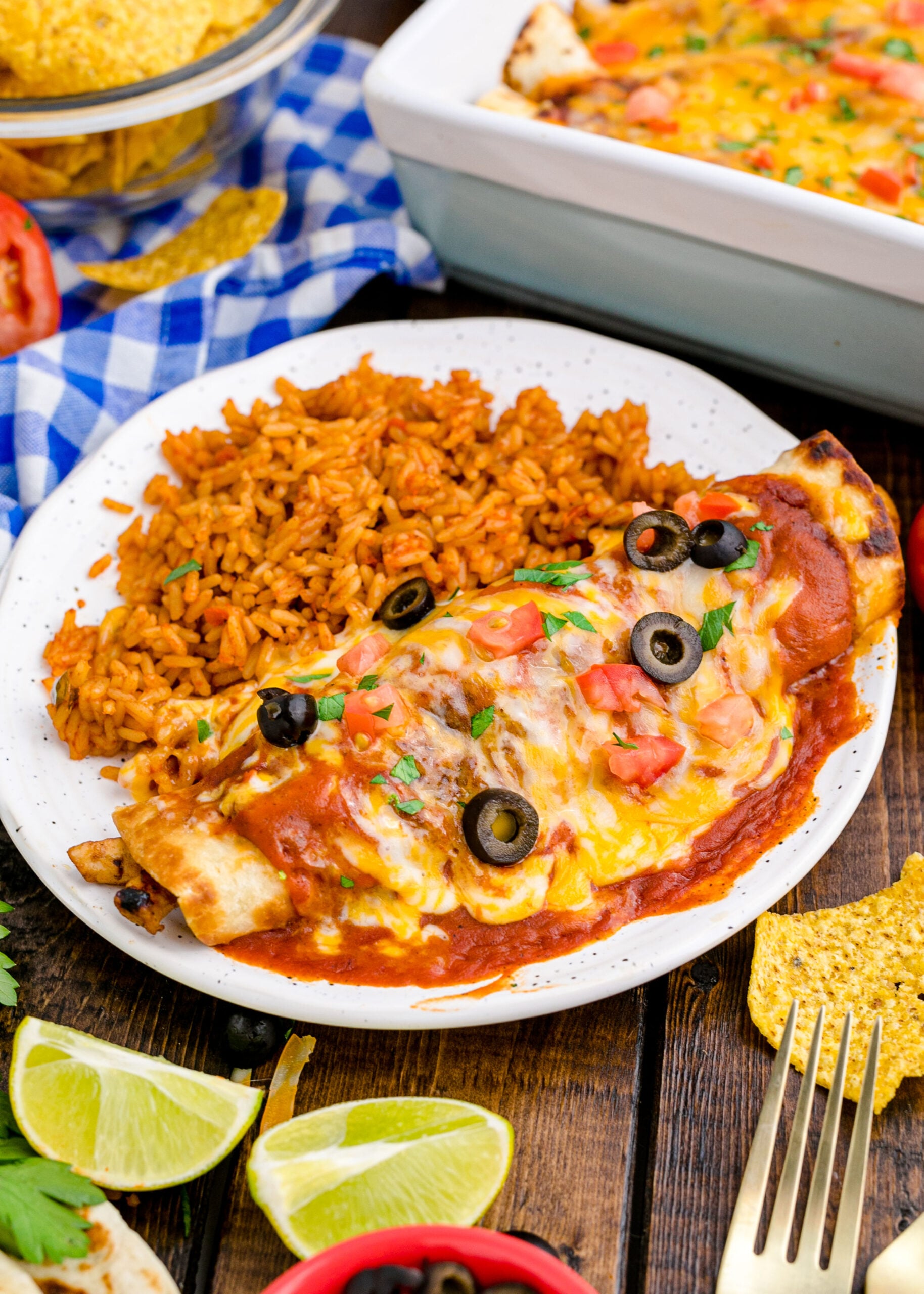 cheesy enchilada on plate covered in sauce and cheese topped with tomatoes, olives, and rice to side
