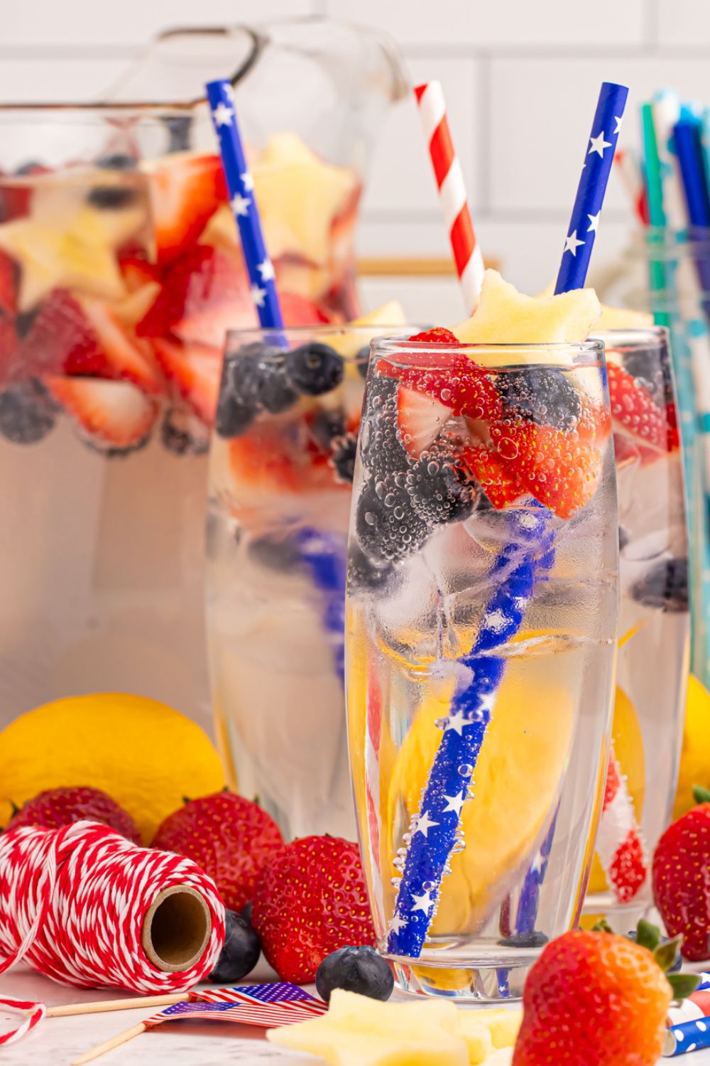 sparkling lemonade in glasses with a pitcher full of lemonade behind it on table there is red and white string on table to add a pop of color 