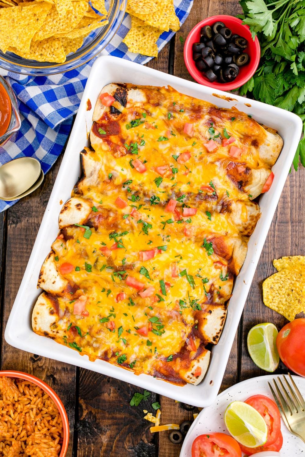 overhead picture of a casserole dish full of enchiladas on table. Condiments and ingredients in bowls on table around the pan 