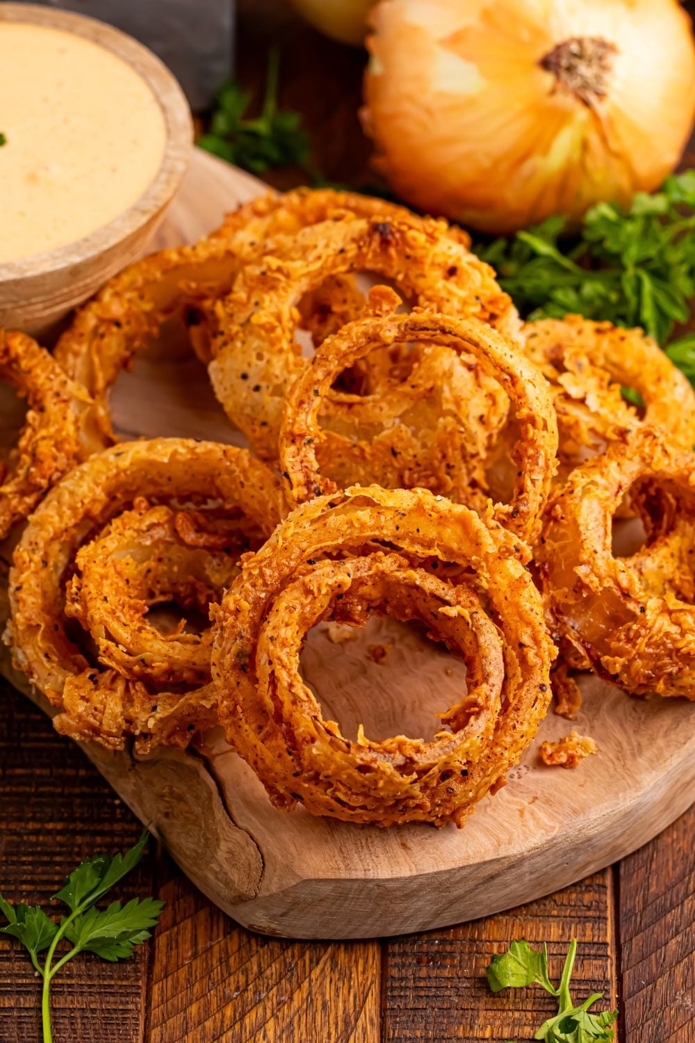 Homemade Deep Fried Onion Rings on a cutting tray with parsley in front and side of tray, with some sauce in a small bowl for dipping