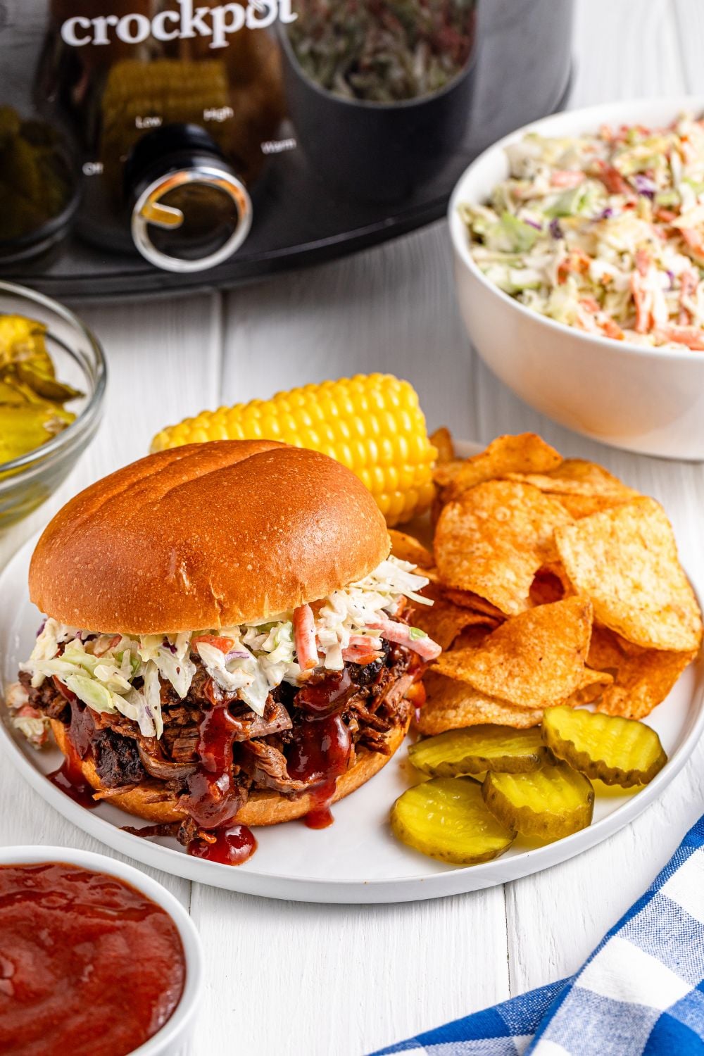 side view of table with bbq brisket with chips, and other items on plate and a slow cooker in background of image