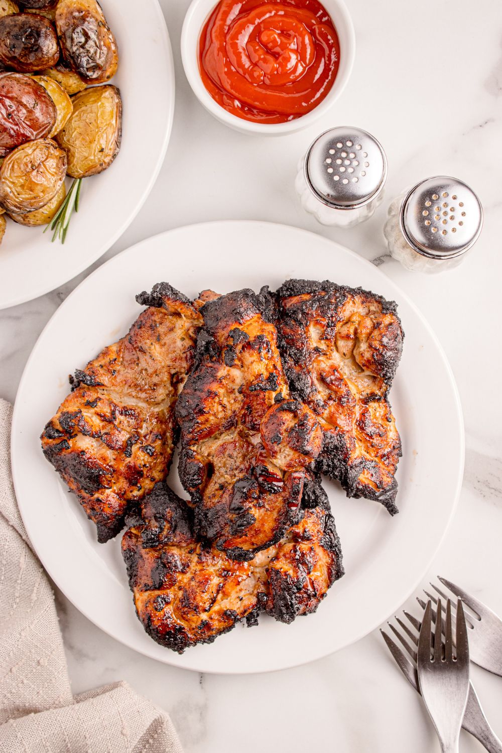 overhead look of Grilled Chicken Thighs on table with salt and pepper, ketchup, and potatoes on platter to the side 