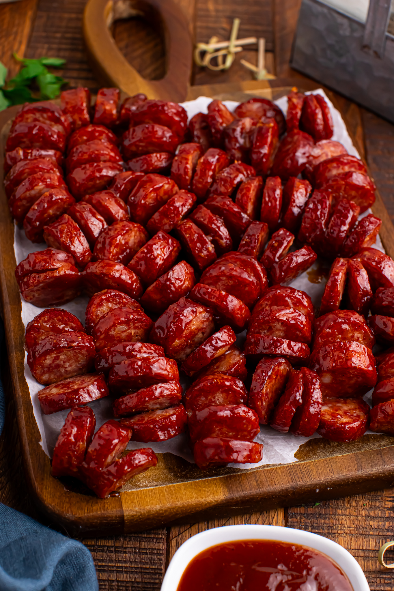 close up view of BBQ Hasselback Smoked Sausages spread out on a dark wood cutting board with a white bowl of bbq in front