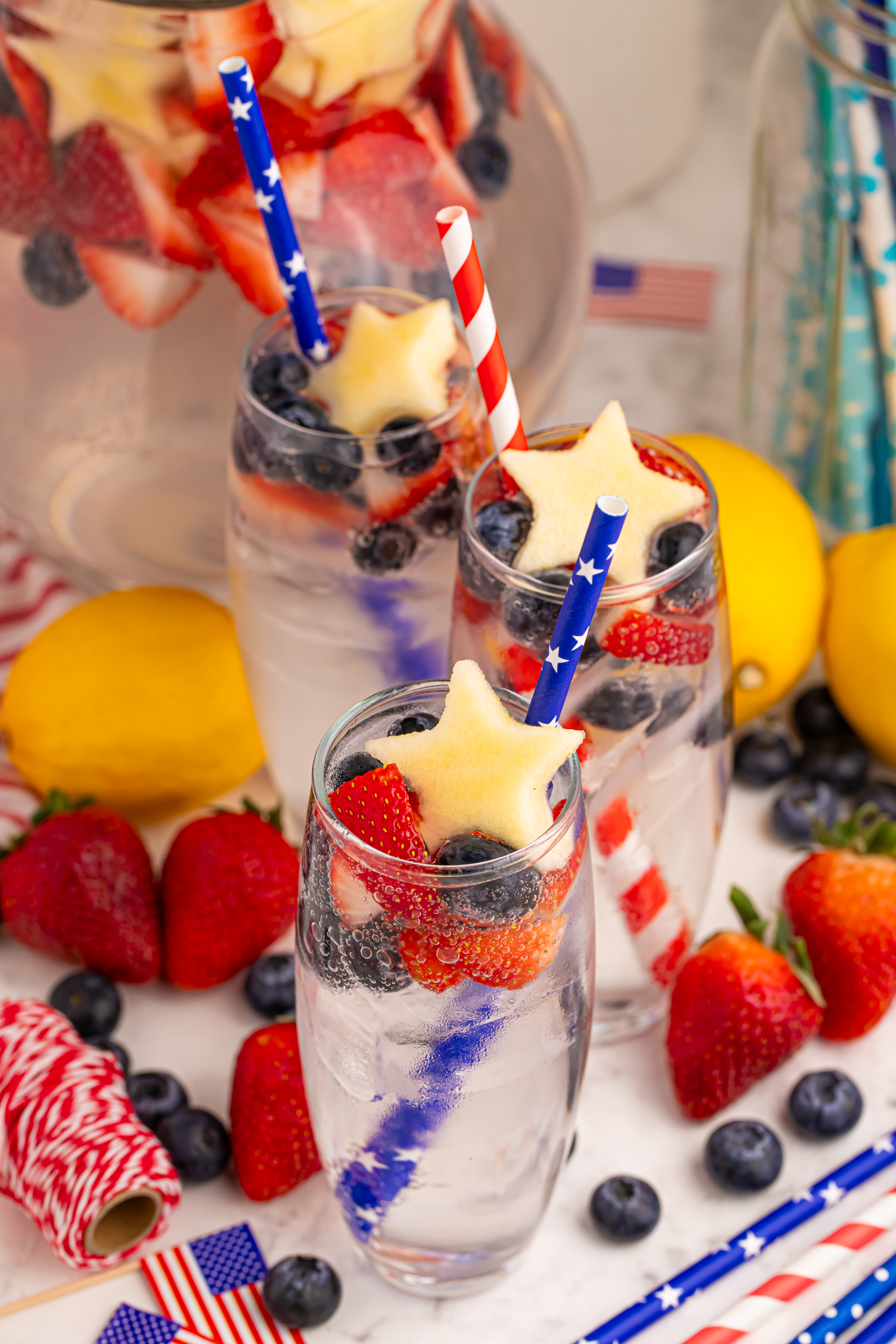 3 tall clear glasses of Patriotic Sparkling Lemonade with flag straws and with fruit scattered on table