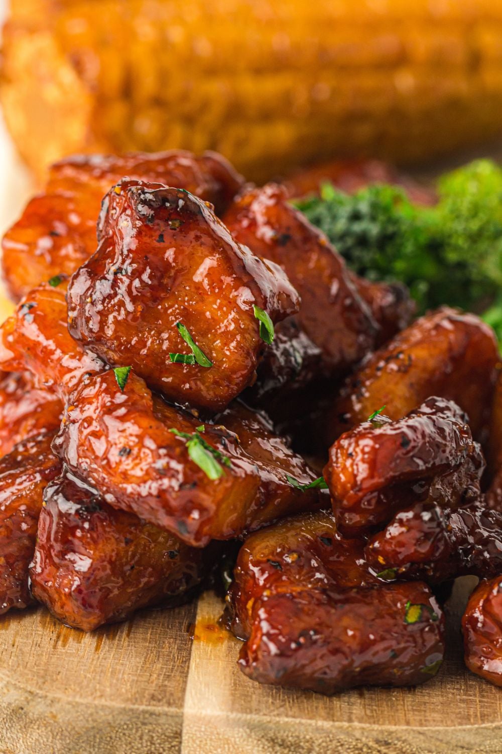Smoked Pork Belly Burnt Ends on a cutting board garnished with herbs and corn on the cob behind it on table