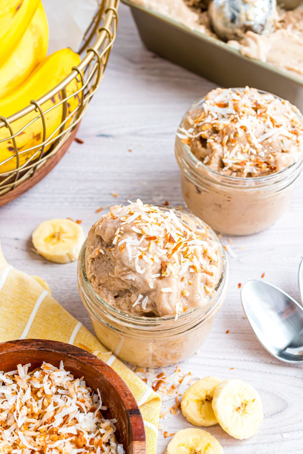 Banana Ice Cream in glass bowls on table with a basket of bananas to side, and spoons and sliced bananas on table 
