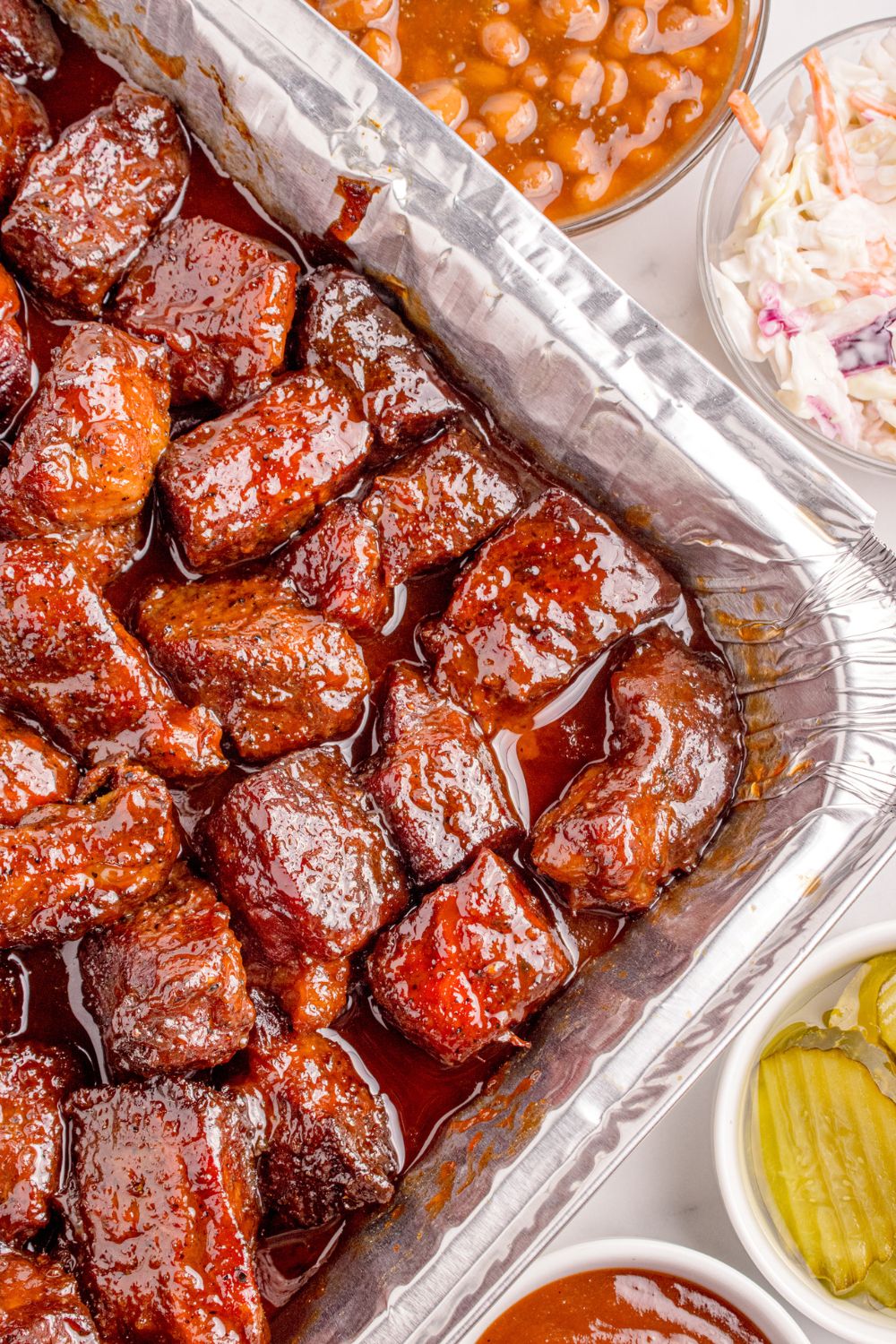 Chuck Roast Burnt Ends in a foil pan on table with beans and coleslaw in bowls to the side of the pan