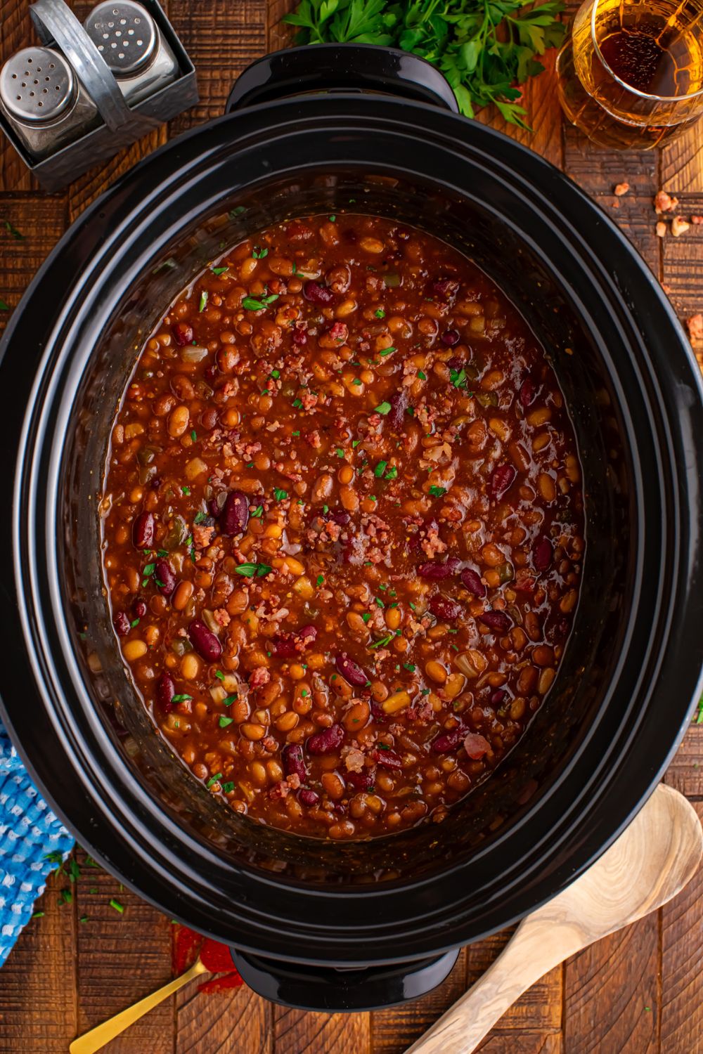 Crockpot bourbon baked beans in crockpot on table with wooden table and garnishes on table