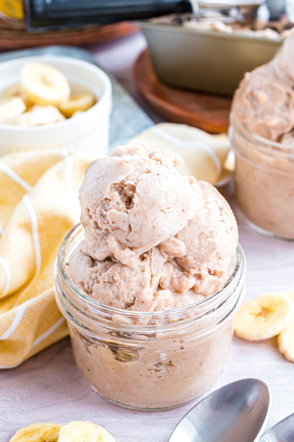 Banana Ice Cream in jars with a yellow towel between bowls and sliced bananas in a bowl behind the ice cream 