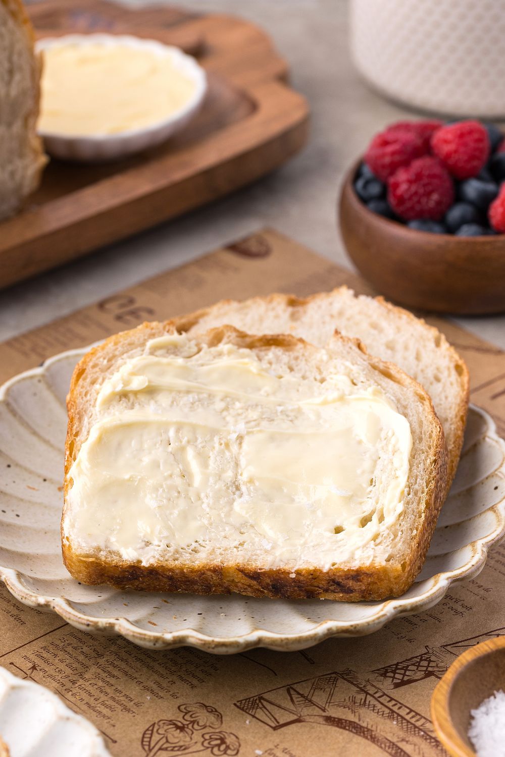 Homemade Sourdough Bread sliced and buttered sitting on plate with a bowl of fruit behind it on table 