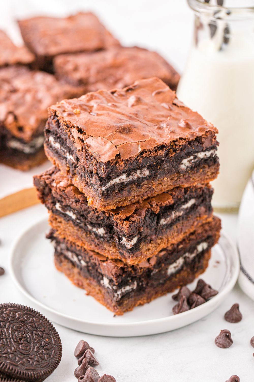Slutty Brownies stacked on a plate with chocolate chips and cookies on table around the bars 