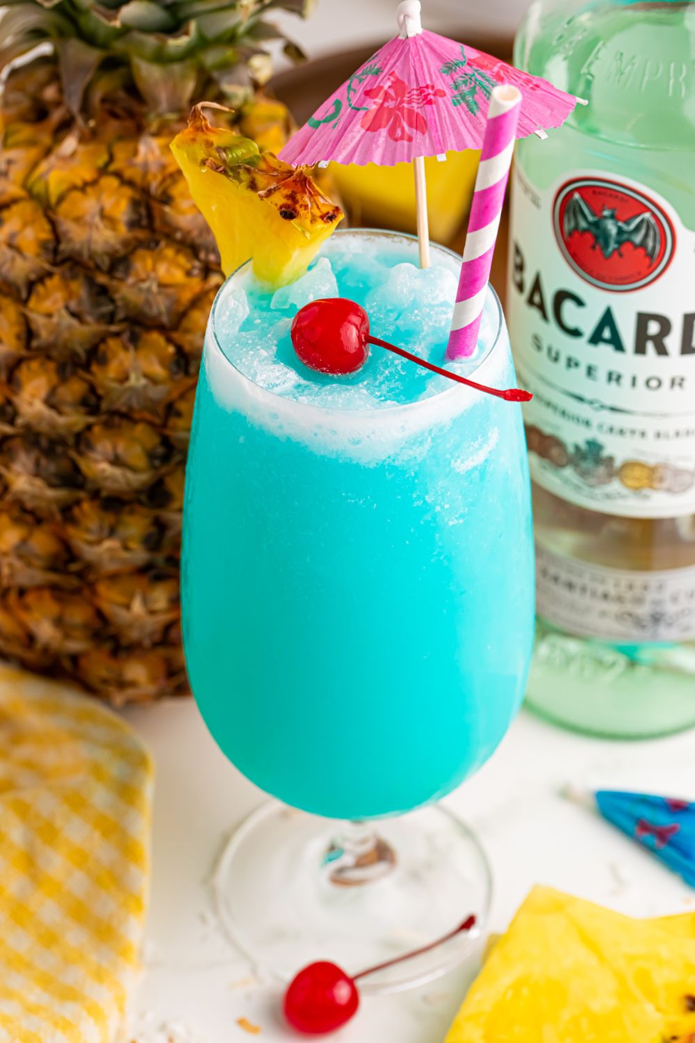 pineapple and bacardi rum on table with a glass of Frozen Blue Hawaiian Cocktail in it and a cherry and pineapple wedge in front of it 