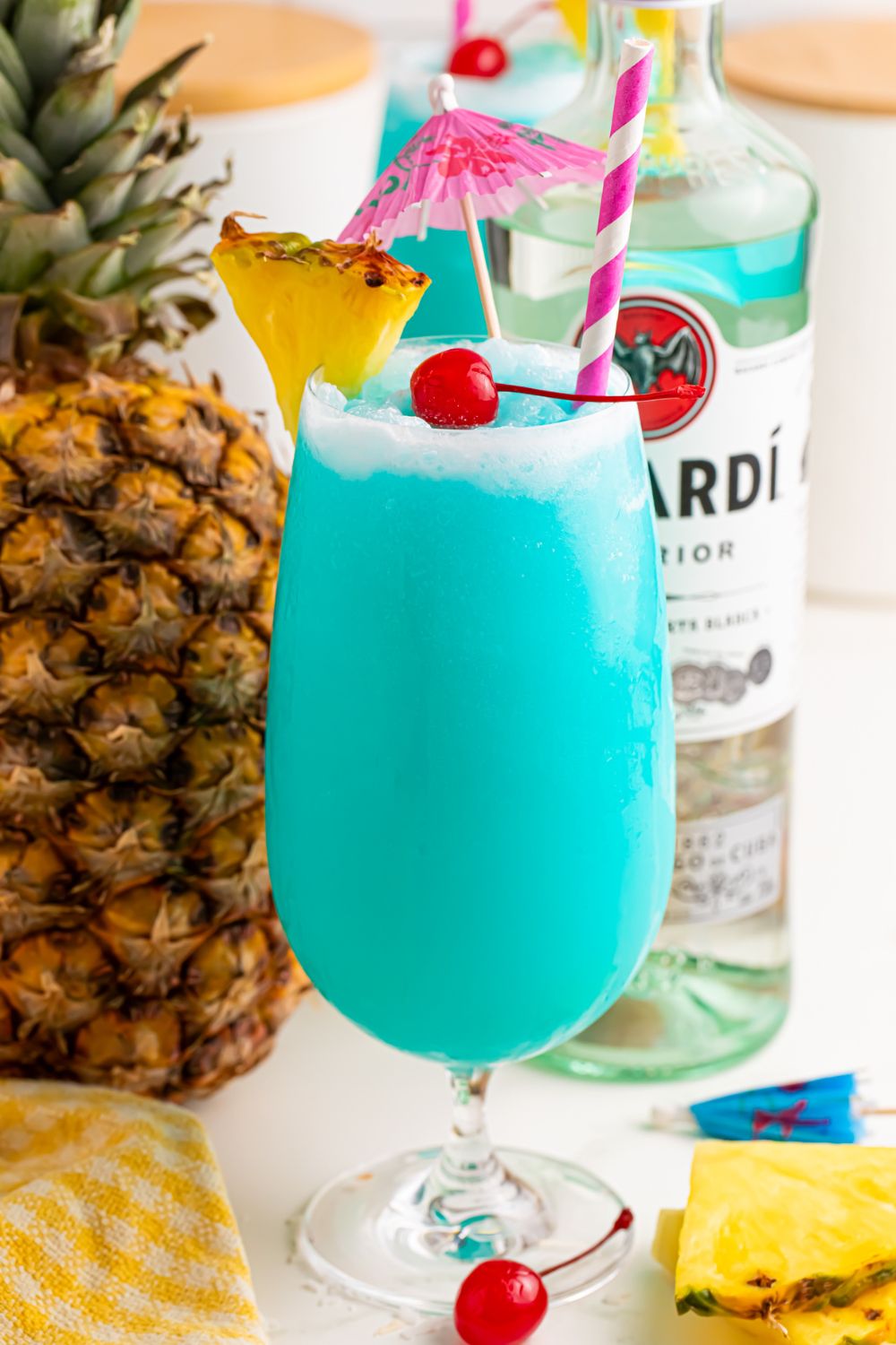 view of Frozen Blue Hawaiian Cocktail with umbrella and straw in glass sitting on a table with ingredients used in it scattered on table 