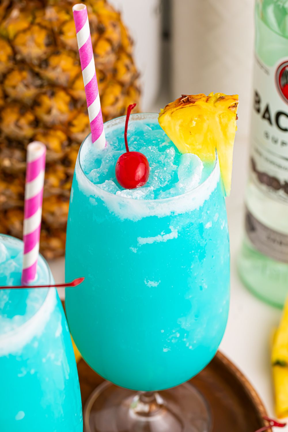 view of blue cocktails in glass with a pineapple on side and cherry in center of glass and pineapple and rum in background
