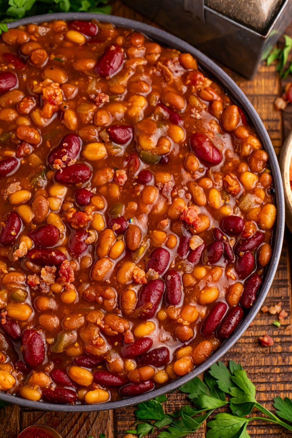 Crockpot bourbon baked beans in a bowl with some herbs around the bowl and salt and pepper