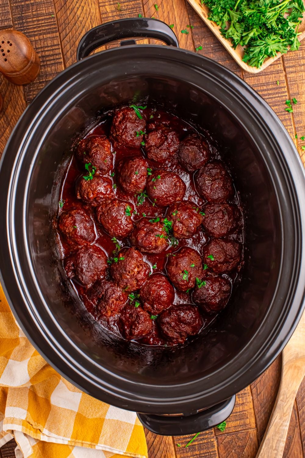 CrockPot sitting on table full of homemade cooked meatballs, with a twoel and garnish to the side of CrockPot