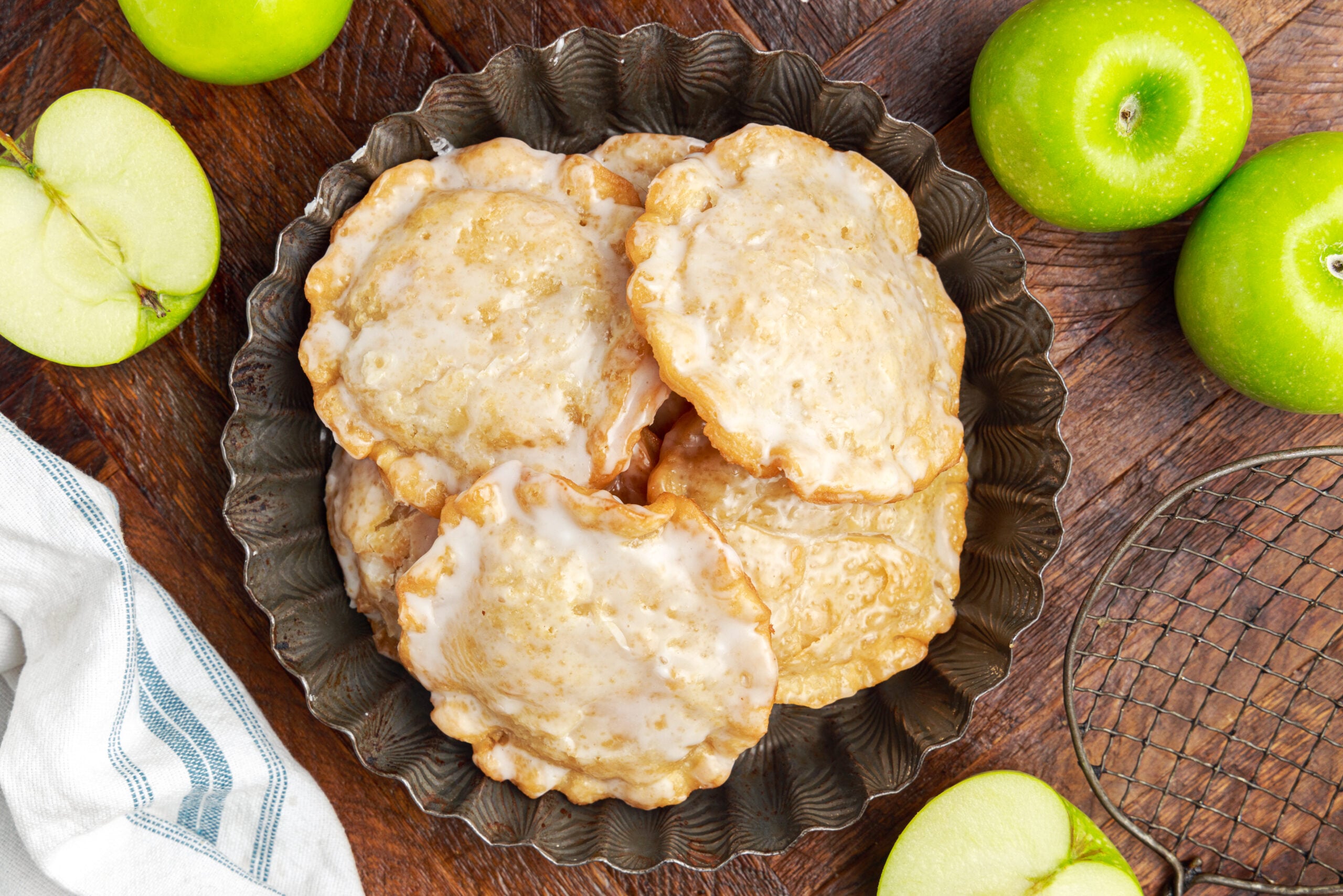 Deep Fried Apple Hand Pies spread around table with apples to side and a bowl of glaze to side