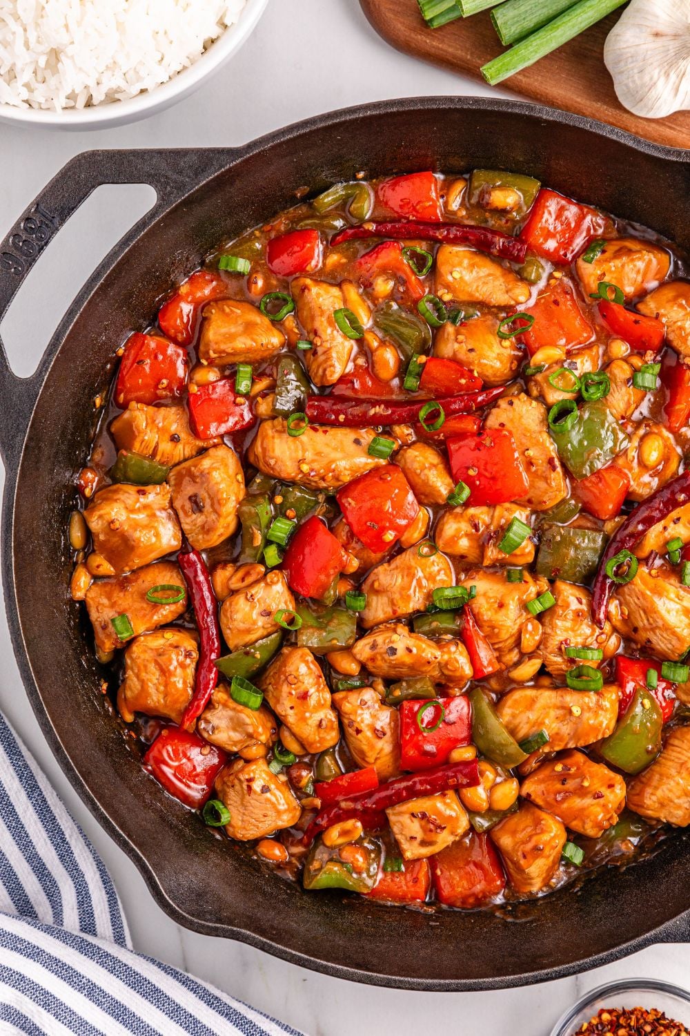 Better Than Takeout Kung Pao Chicken in a skillet on table with green onions and garlic to side