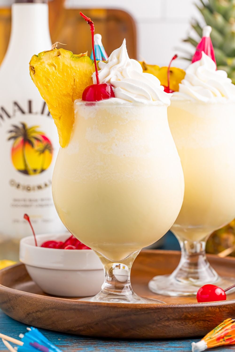 two Frozen Malibu Piña Coladas sitting on a tray with bowl of cherries, and a bottle of Malibu rum to the side 