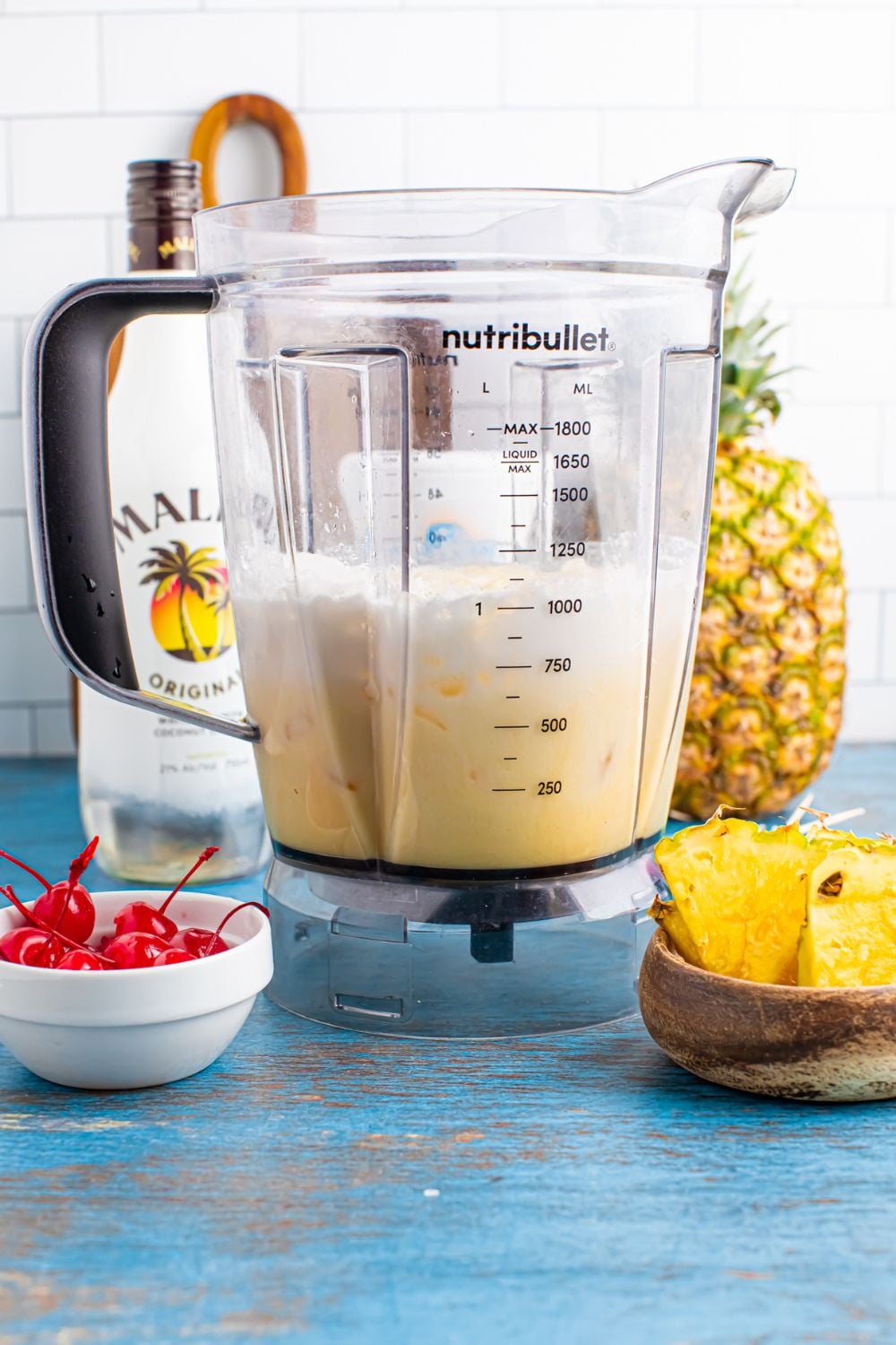 half mixed Frozen Malibu Piña Colada with a bowl of cherries and a bowl of pineapple, and Malibu rum in a bottle on table