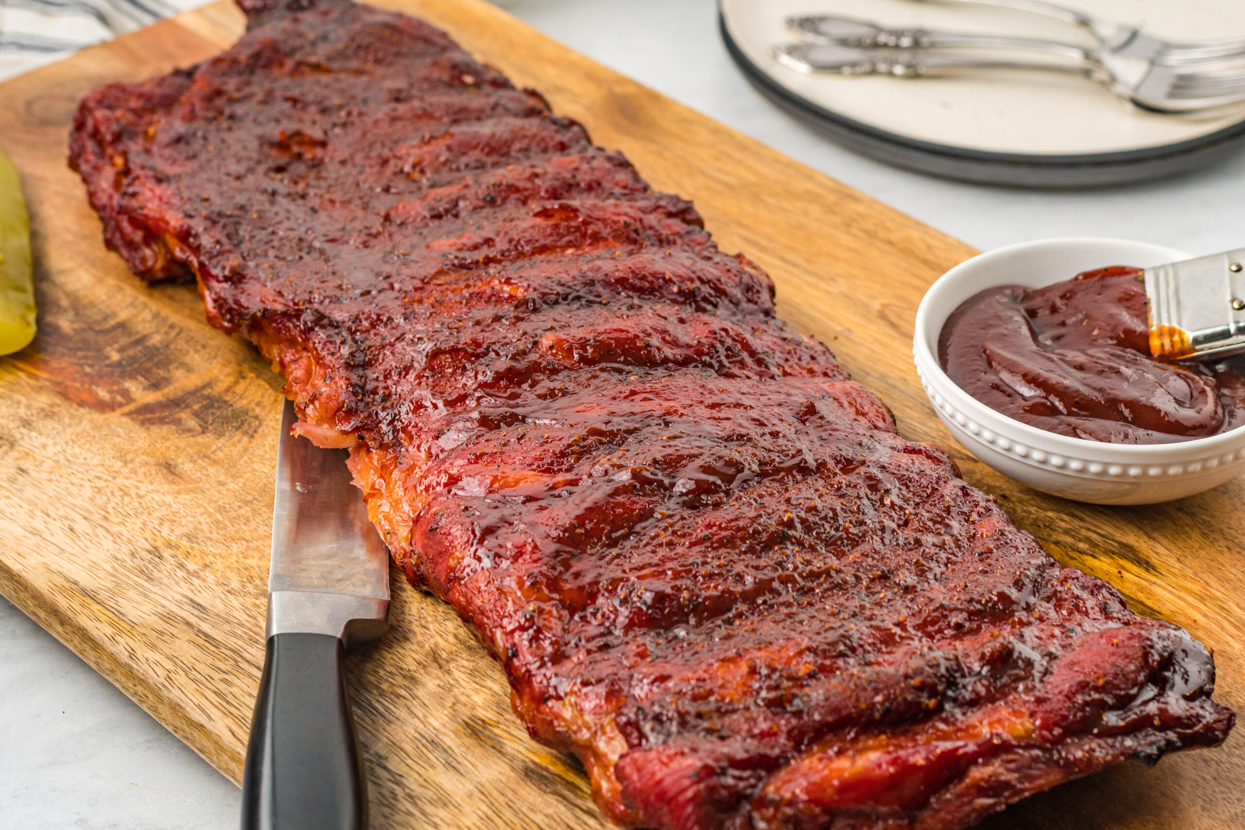 Grilled BBQ Baby Back Ribs on a wooden cutting board with more sauce in a bowl next to them