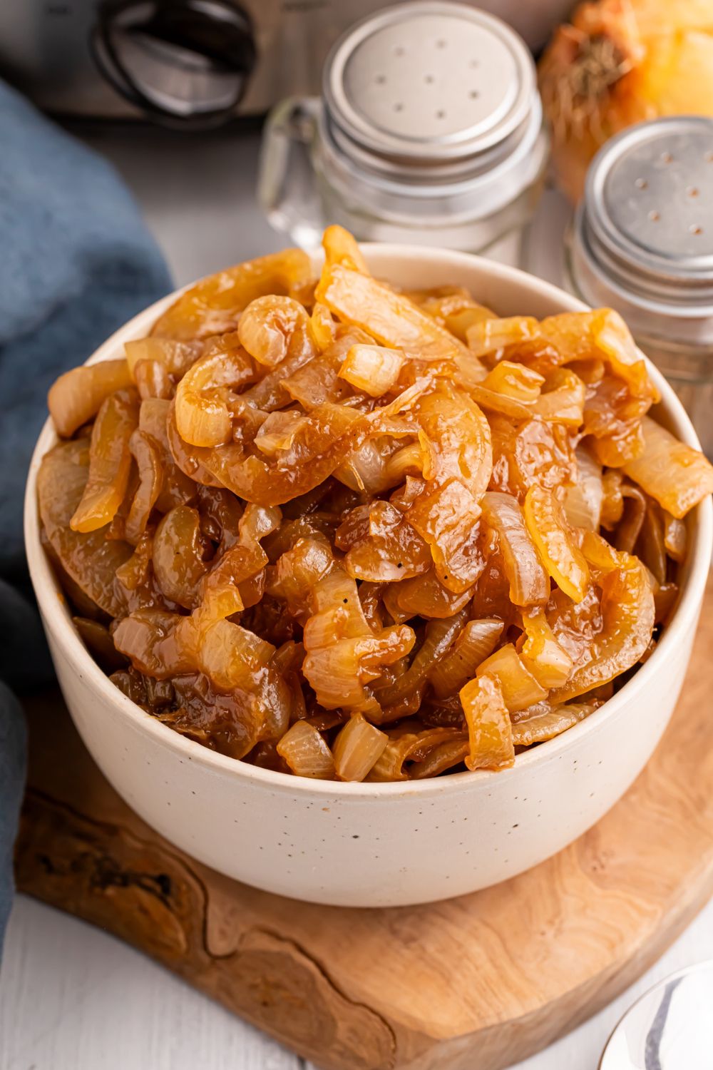 CrockPot Caramelized Onions in a bowl on a cutting board on the table. Salt, pepper, and onion to the side of the container