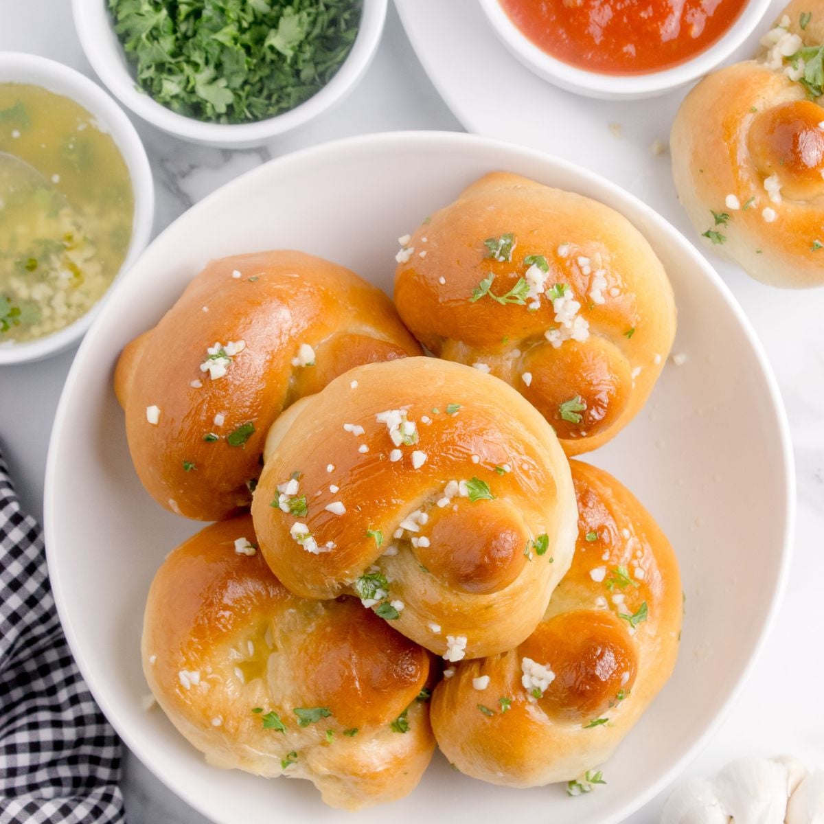 Homemade Garlic Knots piled out on a plate with butter, parsley and dipping sauces to side