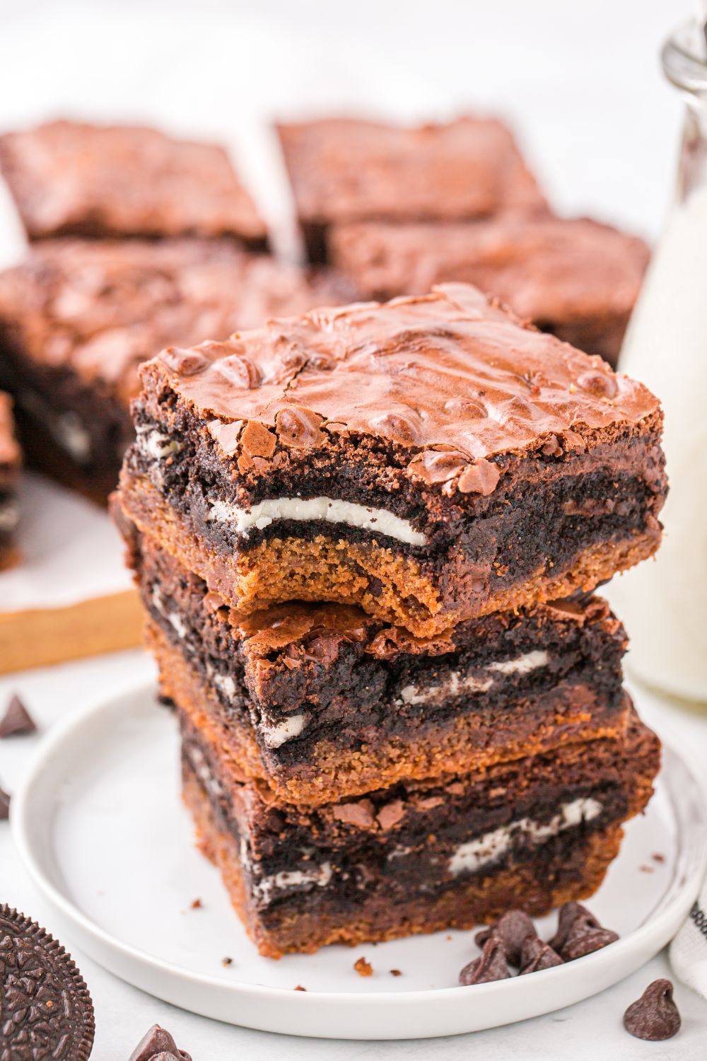 stack of Slutty Brownies on a white plate with a bite taken out of one, and behind a tray with the rest of the bars laid out 