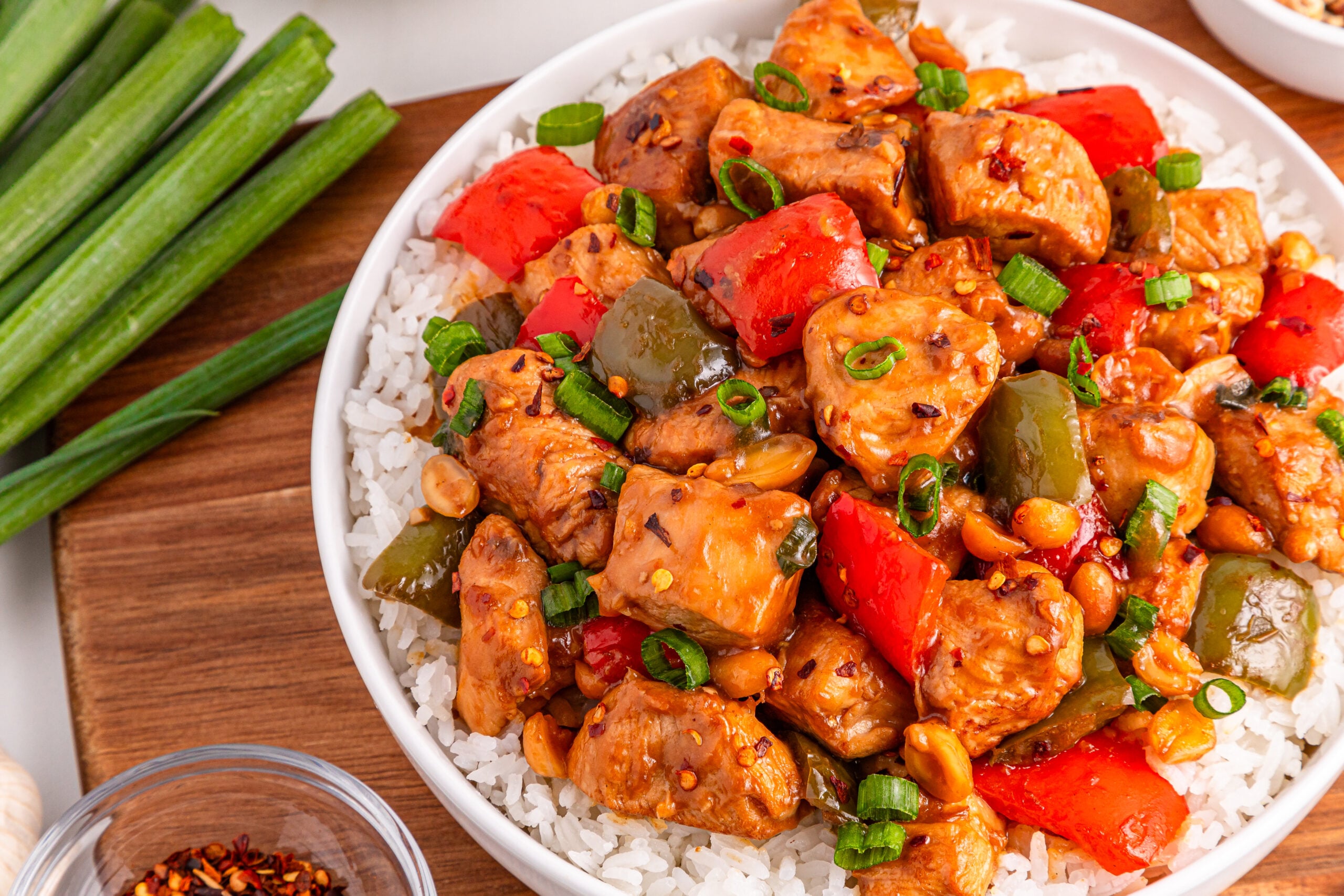 Better Than Takeout Kung Pao Chicken in a bowl with a bed of rice and cutting board with vegetables to the side