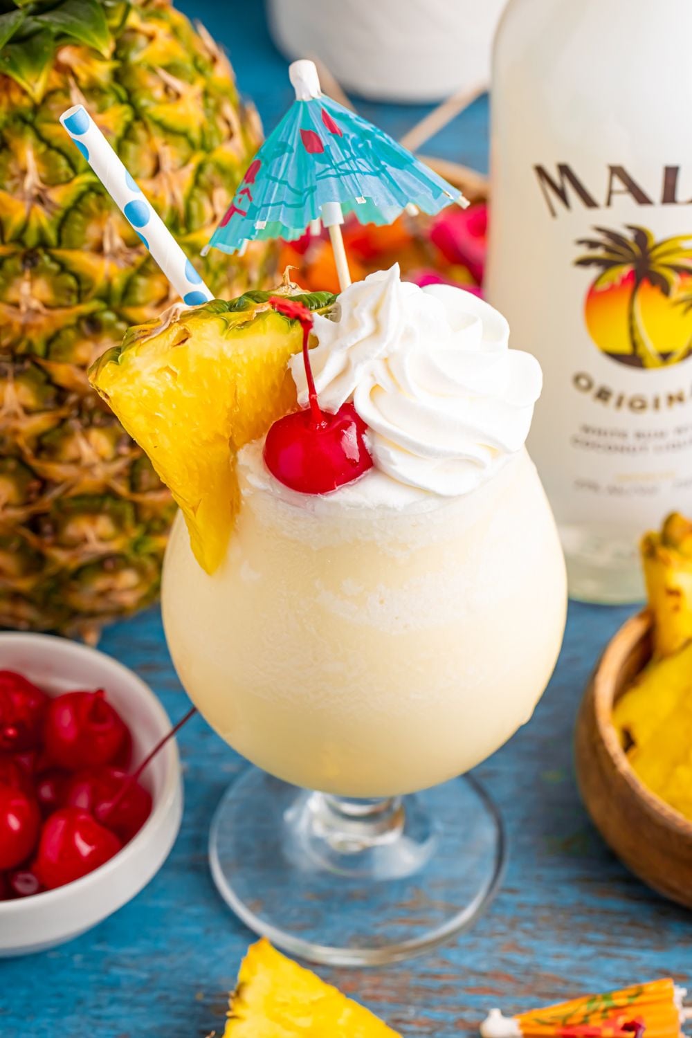 Frozen Malibu Piña Colada on table with whole pineapple and bottle of rum on table as well 
