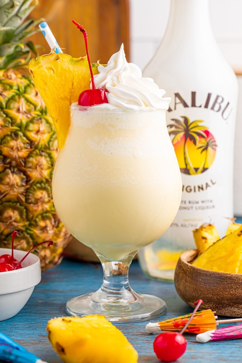 Frozen Malibu Piña Colada in a glass on table with pineapple slices and malibu and a whole pineapple to side on table 