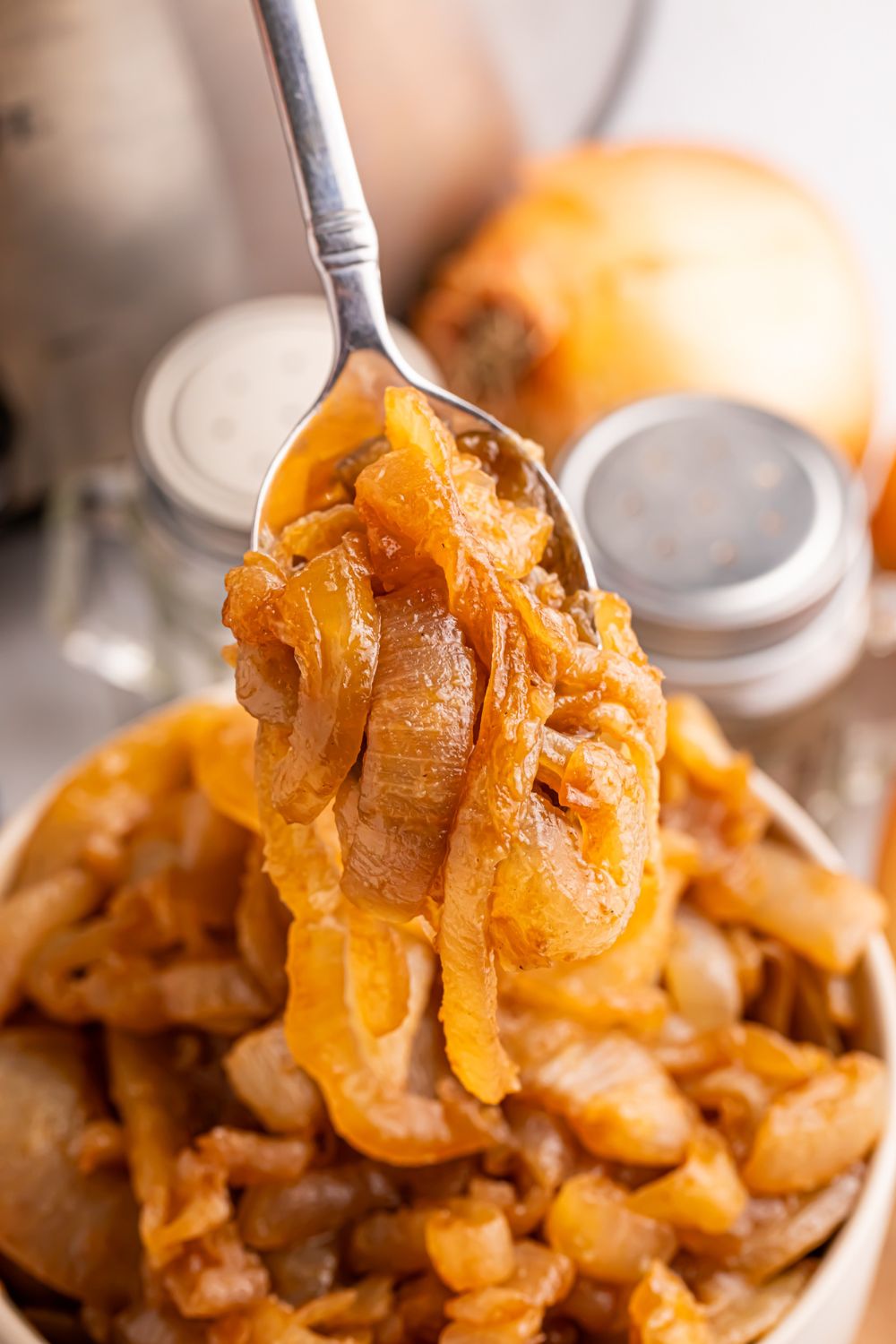 spoon scooping up some of the CrockPot Caramelized Onions out of a bowl on table. Salt and pepper shakers to the side of bowl on table