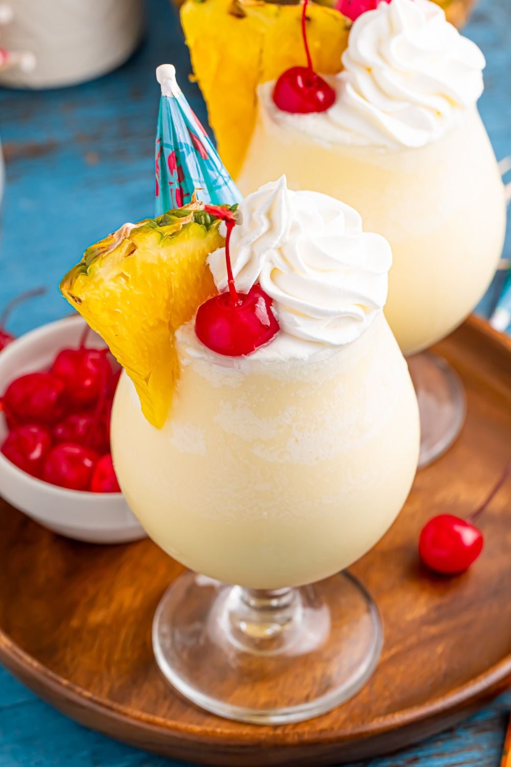 Frozen Malibu Piña Colada glasses on table with a small cocktail bowl of cherries to the side all on a tray 