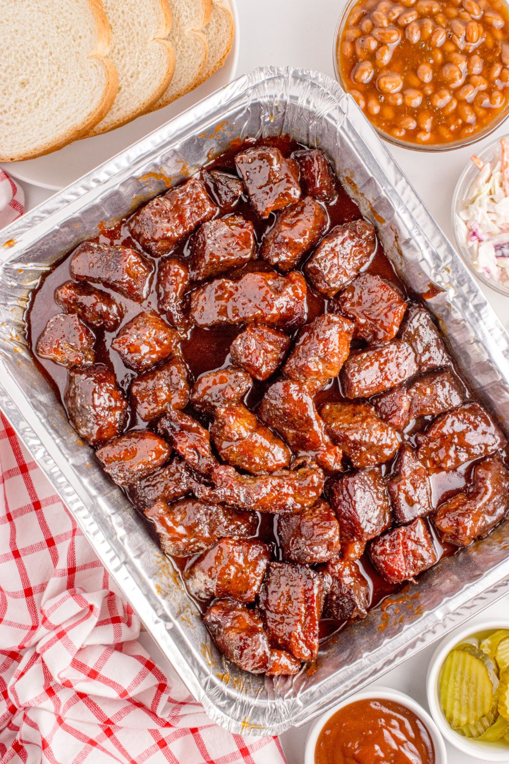 Chuck Roast Burnt Ends in a foil baking pan on table with bread, beans, ketchup, and pickles in bowls around table