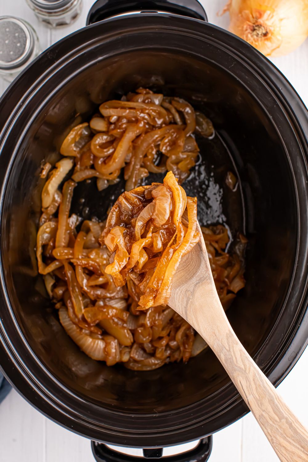 Crockpot with Caramelized onions in it with a wooden spoon stirring some of the onions around