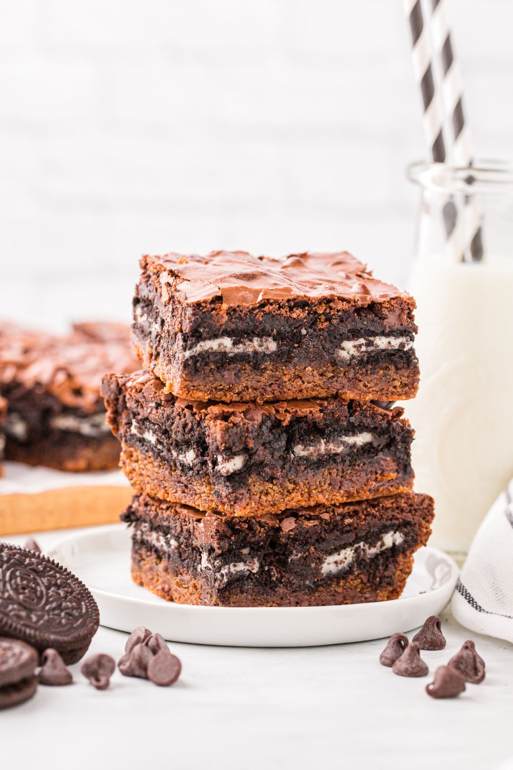 Slutty Brownies stacked on table with chocolate chips and Oreo cookies to the side and a glass of milk with straws in it 