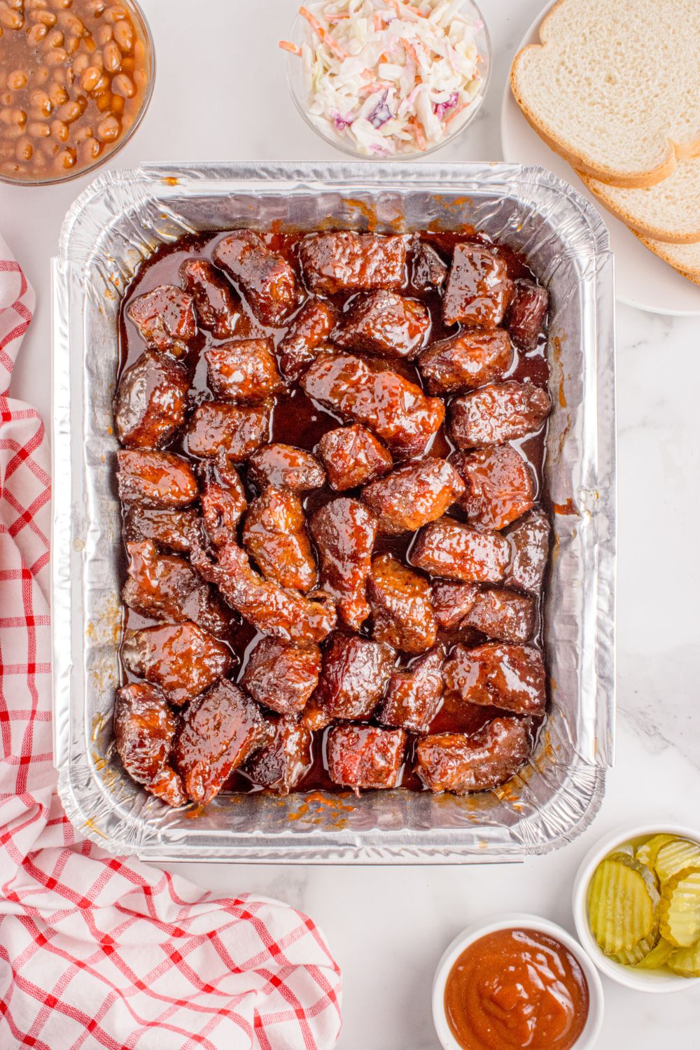 Chuck Roast Burnt Ends in a tin foil pan with bread, coleslaw, pickles, and more all around the pan