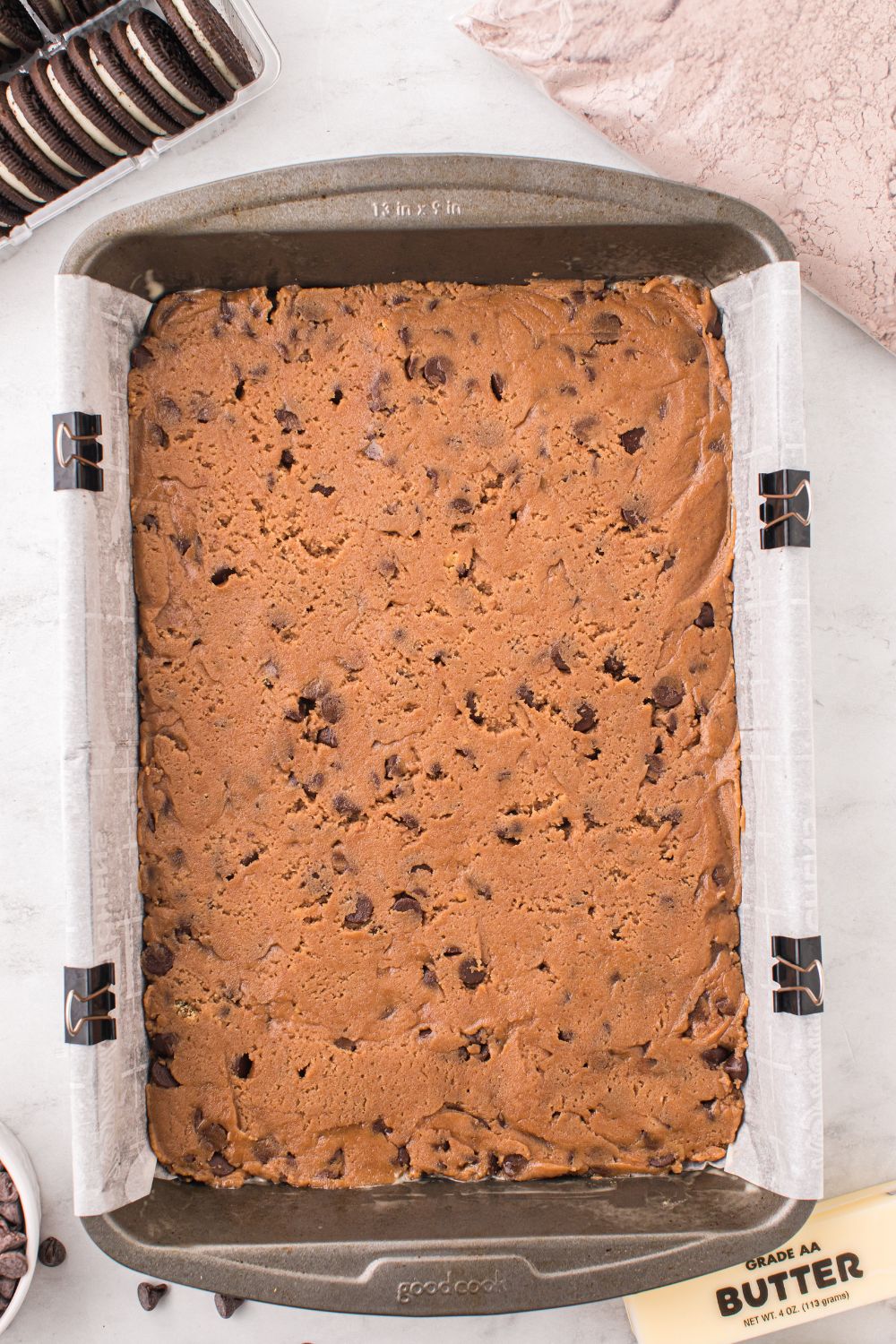 cookie dough batter in a 9x13 pan on table with Oreo cookies to side and a bag of brownie mix 