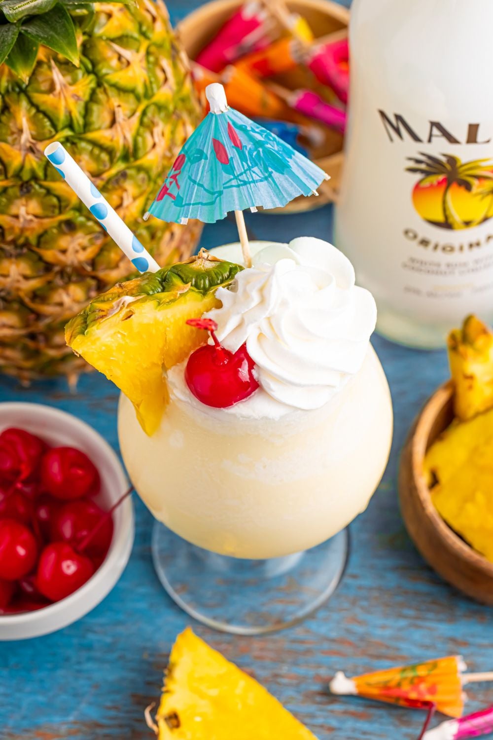Frozen Malibu Piña Colada on table with a pineapple in the background and a bowl of cherries to side, and bottle of Malibu rum