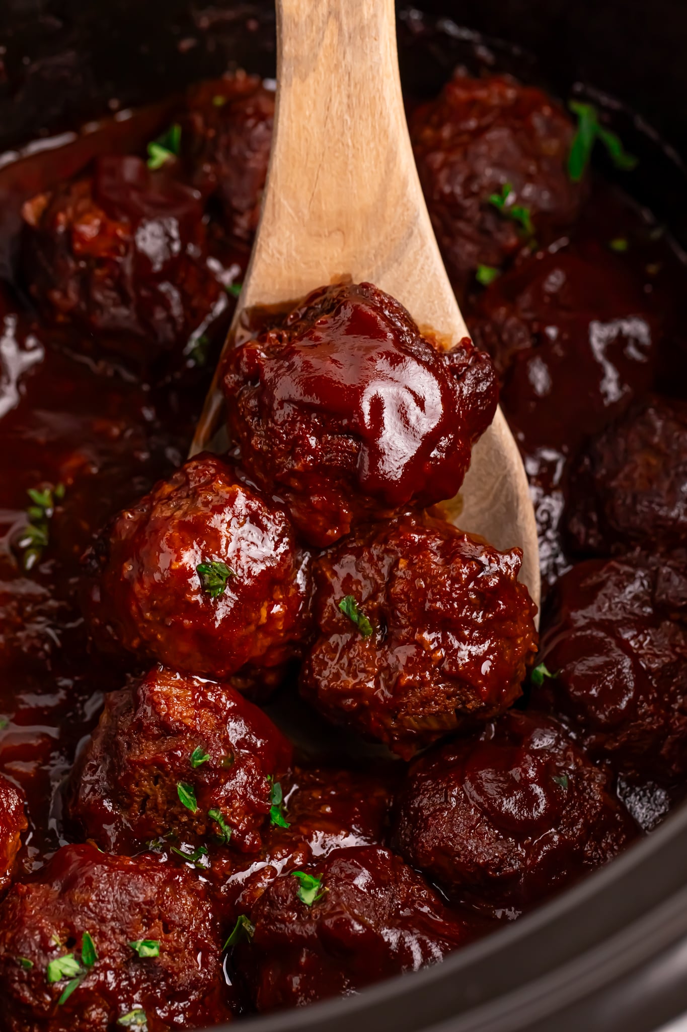 Crockpot Homemade BBQ Meatballs in a black slow cooker with a wooden spoon holding 3 up
