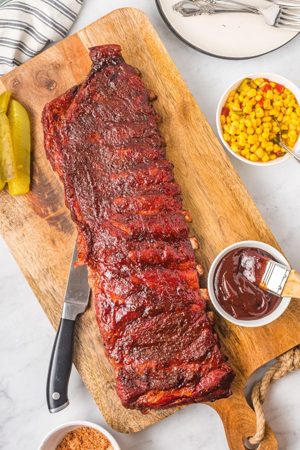 Grilled BBQ Baby Back Ribs on cutting board with sauce and corn to side in bowls