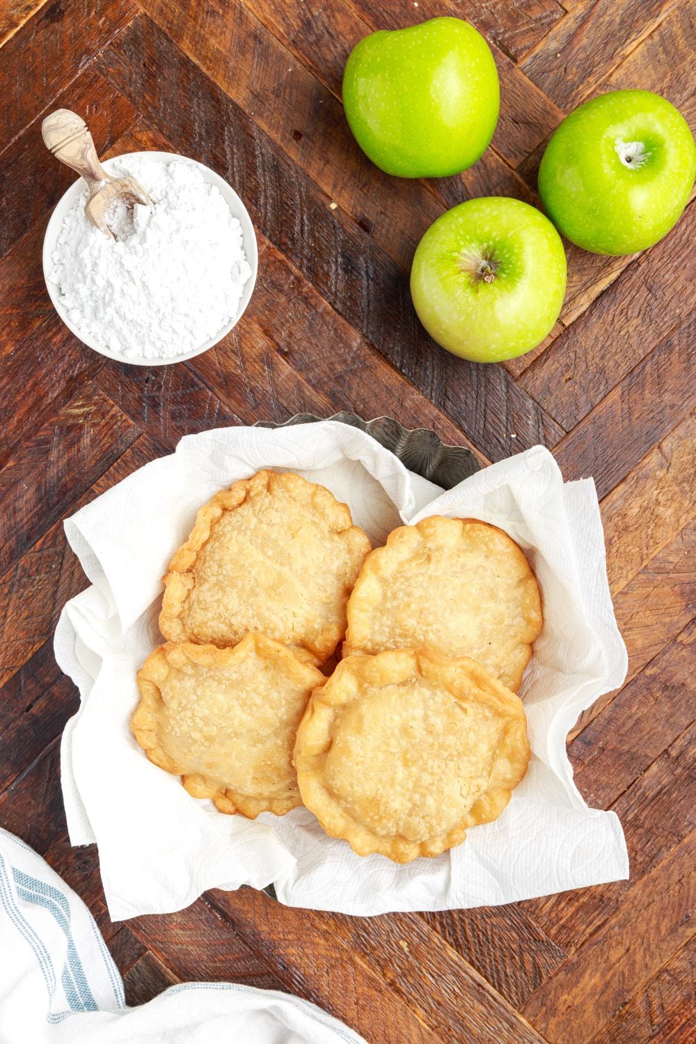 Deep Fried Apple Hand Pies in a basket on table with powdered sugar to the side and green apples